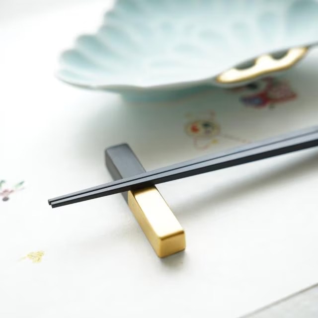 Black chopsticks resting on a matte black and gold Arita cutlery rest beside a pale blue plate