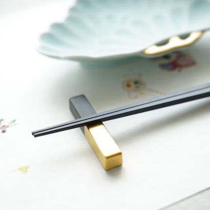 Black chopsticks resting on a matte black and gold Arita cutlery rest beside a pale blue plate